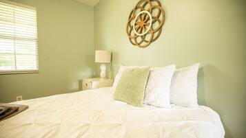 Cama bem arrumada com roupa de cama branca, um objeto decorativo na parede e uma luminária de cabeceira.