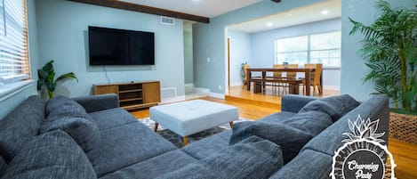A living room with grey sofas, a flat-screen TV, wooden beams on the ceiling, and a dining area with a table and chairs.