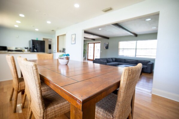 A spacious dining area with a wooden table and wicker chairs, a kitchen in the background, and a living area with a sofa and large windows.