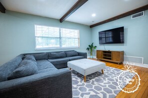 Un salón con un sofá seccional gris, un reposapiés blanco, una alfombra estampada, un mueble de entretenimiento de madera y un televisor de pantalla plana.