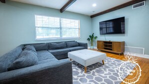 A living room with a grey sectional sofa, a white ottoman, a patterned rug, a wooden entertainment unit, and a flat-screen TV. - Winter Park Charmer - Heated Pool Home - Close to everything (Winter Park)