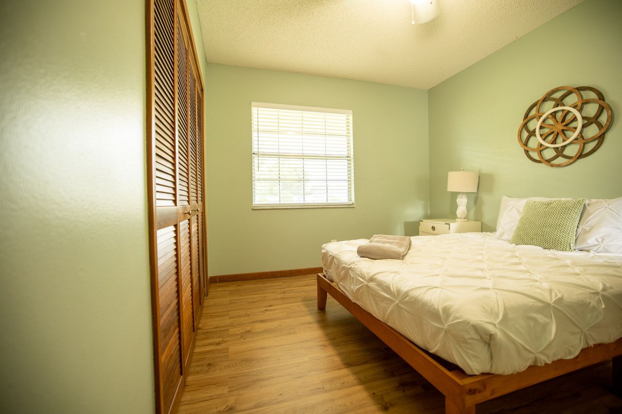 Quarto com cama de madeira, janela com persianas, mesa de cabeceira com luminária e um objeto decorativo na parede.