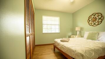 Quarto com cama de madeira, janela com persianas, mesa de cabeceira com luminária e um objeto decorativo na parede.