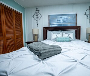 Una cama bien tendida con un edredón doblado, un cabecero de madera, dos mesitas de noche con lámparas y una ventana con persianas.