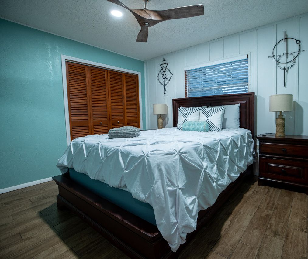 Un dormitorio con cama, cabecero de madera, dos mesitas de noche con lámparas, un ventilador de techo y un adorno en la pared.
