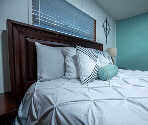 Een net opgemaakt bed met een witte dekbedovertrek en gestreepte kussens, een houten hoofdbord en een muur in blauwgroen.