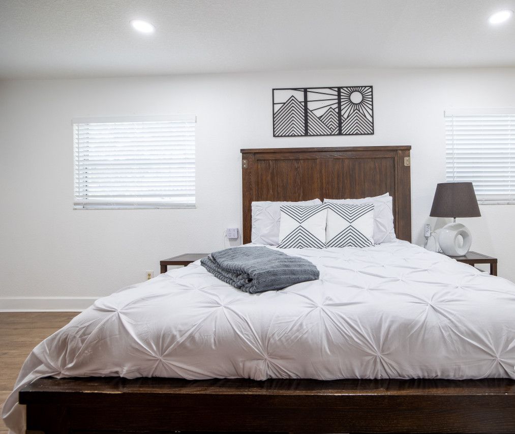 Quarto com cabeceira de madeira, roupa de cama branca, manta cinza, abajur e um objeto decorativo na parede.