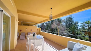 Un balcó amb taula i caires, un sofà amb coixins i una barana amb vista a un jardí ple d'arbres.