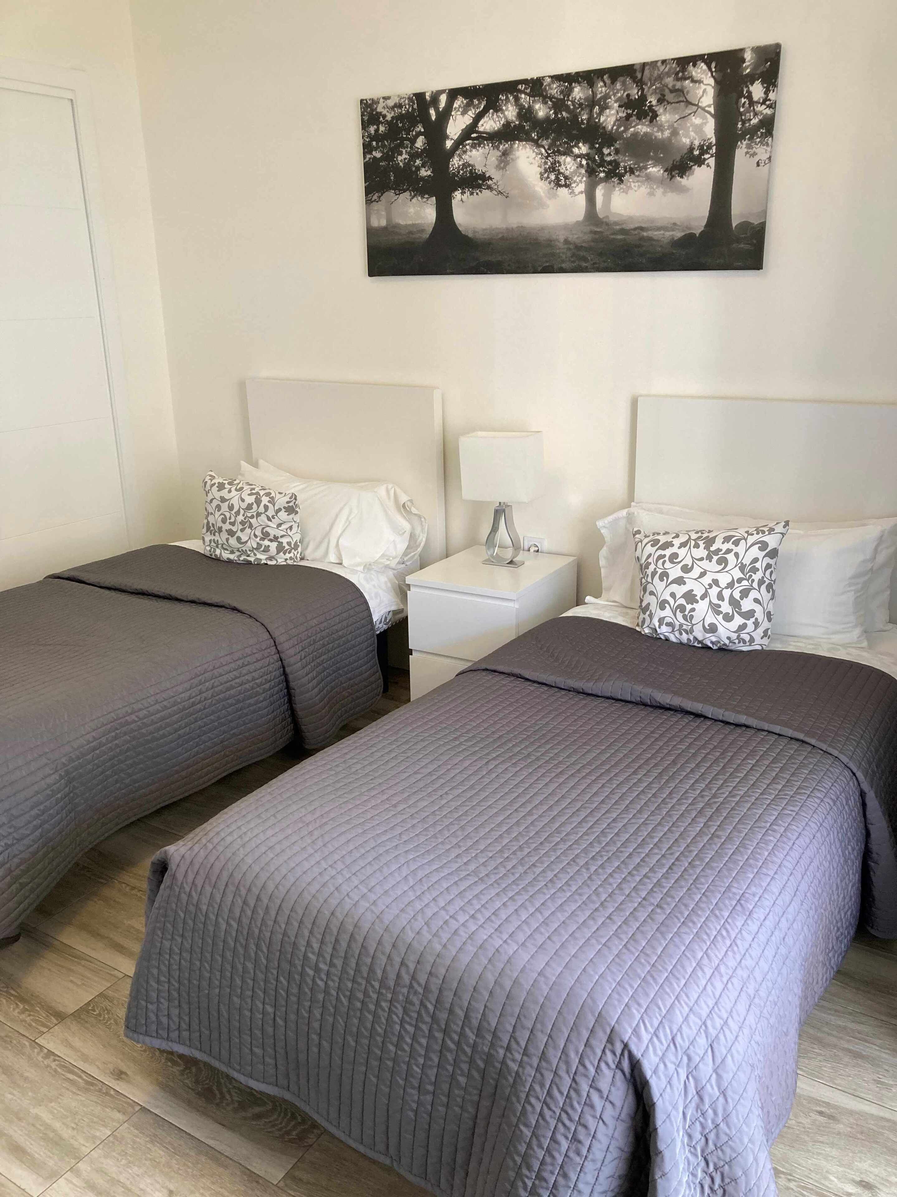 A hotel room with two beds, each with a grey quilted bedspread and matching pillows. There is a white nightstand with a lamp between the beds. A large black and white tree painting hangs on the wall above the beds.
