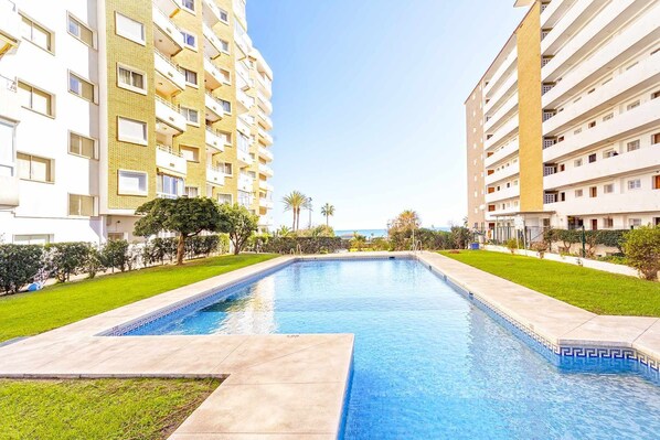 A swimming pool surrounded by well-maintained grass and trees, with high-rise buildings in the background. - Club Maritimo Front line Beach Apartments (Fuengirola)
