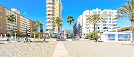 A beachfront promenade with modern buildings, palm trees, and a clear blue sky.