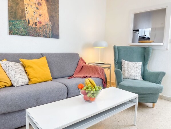 Un salon avec un canapé gris, un fauteuil vert, une table basse blanche sur laquelle se trouve un panier de fruits, et un tableau accroché au mur.