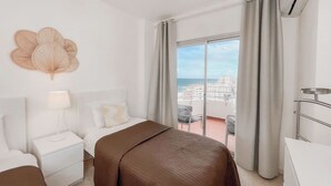 A hotel room with a bed, a nightstand, a lamp, a view of the city and beach through a large window, and an air conditioning unit. - Club Maritimo Front line Beach Apartments (Fuengirola)