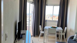 Um quarto pequeno e bem iluminado, com ventilador de teto, uma mesa de jantar com cadeiras, um sofá e uma TV sobre um suporte.