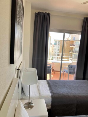 A hotel room with a bed, a bedside table with a lamp, a large window with curtains, and a view of the outside balcony area. - Club Maritimo Front line Beach Apartment (Fuengirola)