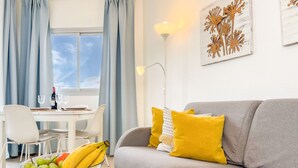A living room with a grey sofa, yellow cushions, a glass table with a fruit bowl, a dining area with a white table and chairs, and a window with curtains.