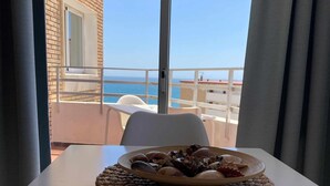 Ein Tisch mit gewebter Unterlage und einer Schale Muscheln, mit Blick auf einen Balkon und freie Sicht aufs Meer.