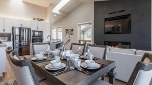 Una cocina moderna con armarios blancos y electrodomésticos de acero inoxidable, un comedor con mesa de madera dispuesta para seis personas y una sala de estar con sofá gris y televisor de pantalla plana montado en la pared.