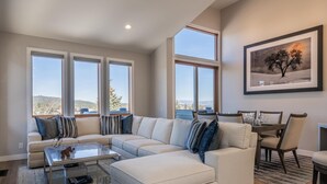 Una sala de estar moderna con un sofá modular, una mesa de centro de vidrio y un comedor con una mesa y sillas. Amplias ventanas ofrecen vistas al exterior.