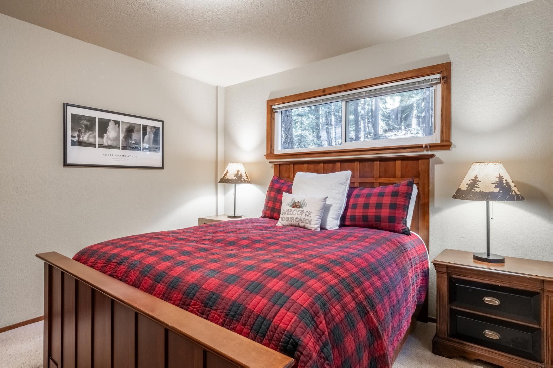 A bedroom with a wooden bed frame, a red and black plaid bedspread, two bedside tables with lamps, a framed picture on the wall, and a window with a view of trees.