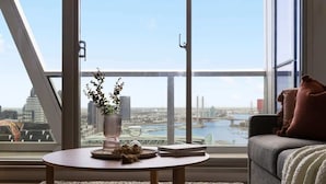 Un soggiorno moderno con un'ampia finestra che offre vista sulla città, un tavolo rotondo in legno con un vaso e oggetti decorativi, e un divano grigio con cuscini.