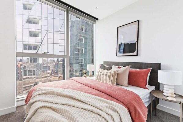 A modern bedroom with a large window, a bed with multiple pillows, a bedside table with a lamp, and a framed picture on the wall.