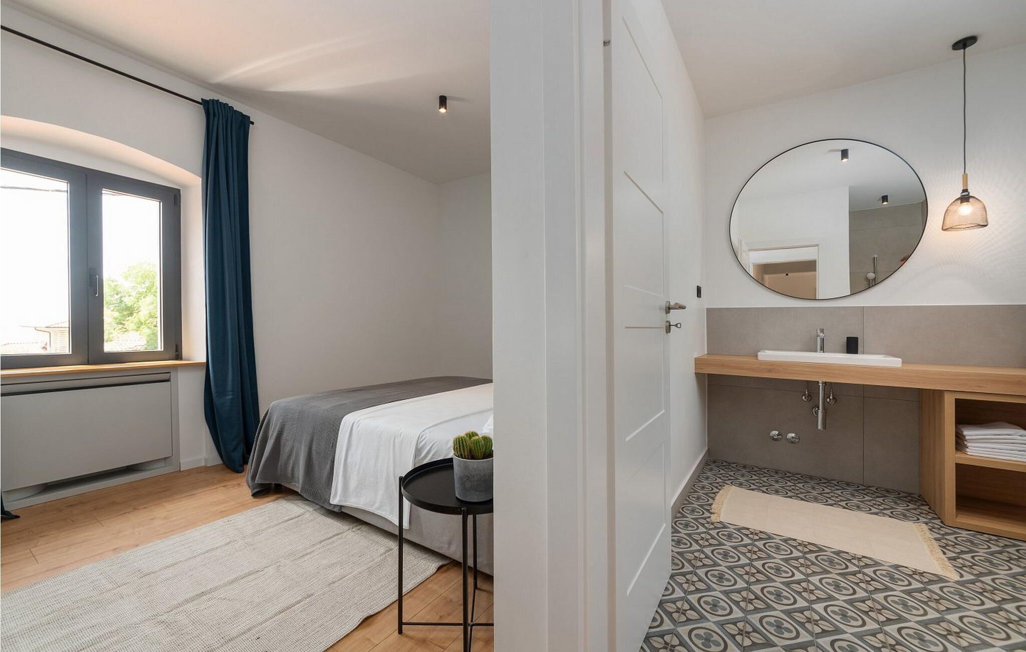 Une chambre à coucher avec un lit, une table de chevet ornée d’une plante, une fenêtre avec des rideaux et une salle de bain équipée d’un lavabo, d’un miroir et d’un carrelage à motifs.