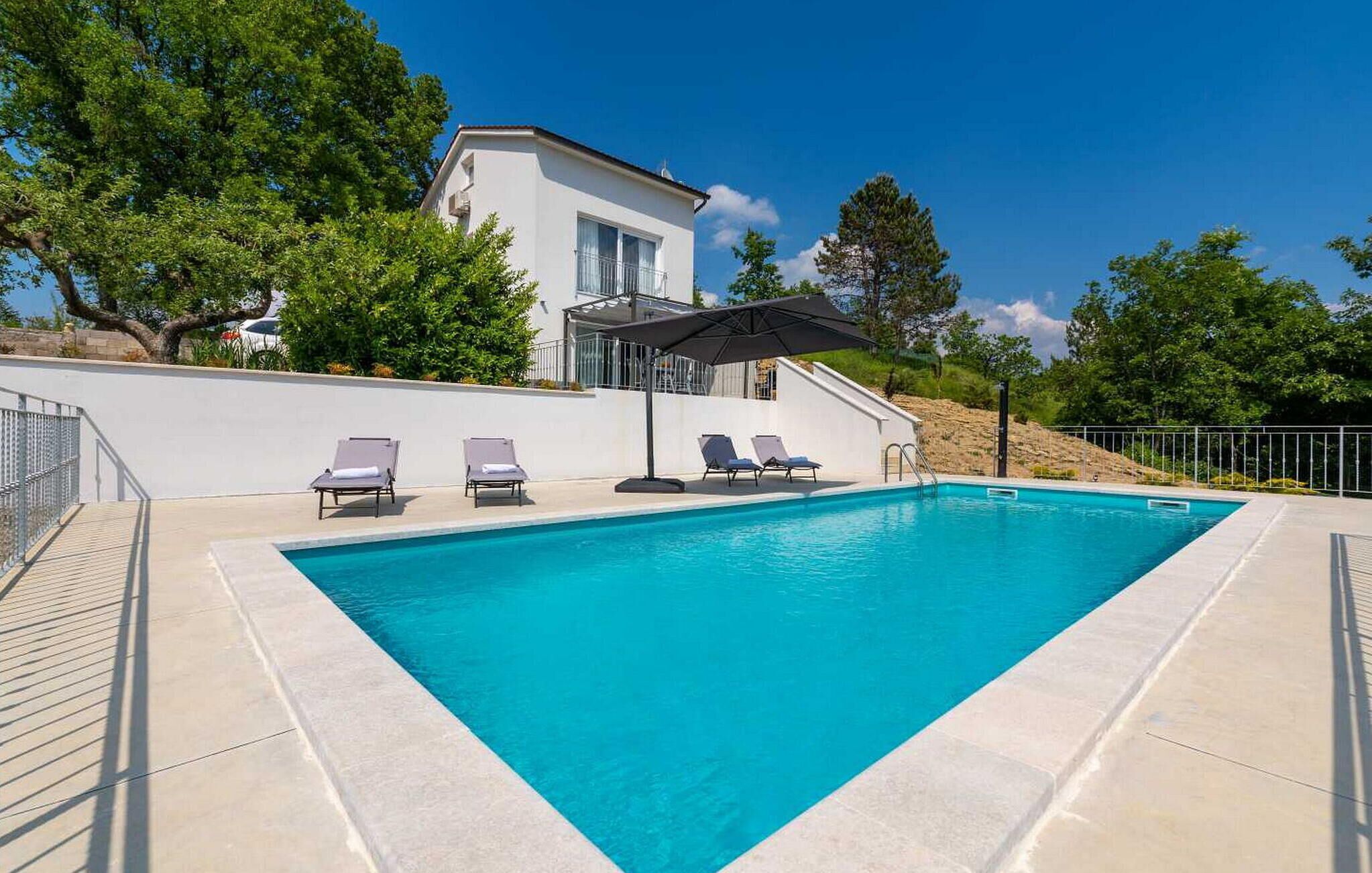 Une piscine rectangulaire bordée d’une zone blanche, avec trois chaises longues et un espace de détente couvert comprenant une table et un parasol.