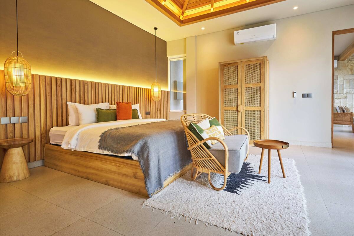 A modern bedroom with a wooden headboard, a large bed with multiple pillows, a wicker chair, a small table, and a white rug.