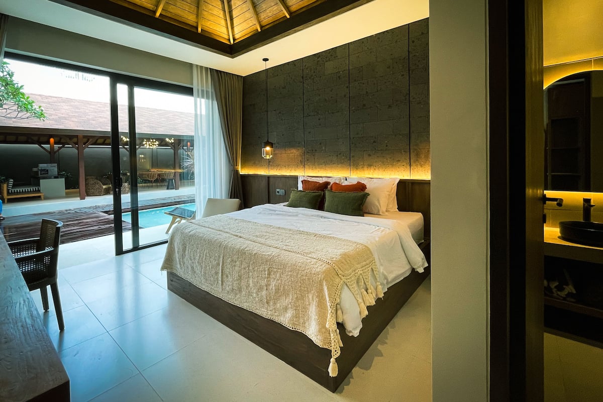 A modern bedroom with a large bed, a view of the outdoors, and a dining area with a pool.