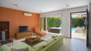 Ein modernes Wohnzimmer mit einer Couch, einem Fernseher, einem Couchtisch und einem Sessel. Eine Glastür führt zu einem Außenbereich mit Pool und Liegestühlen.
