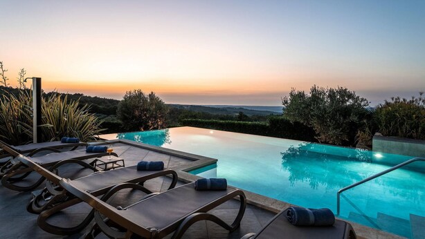 A poolside area with lounge chairs and a glass table, overlooking a clear pool and a scenic view during sunset.