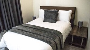A neatly made bed with a dark headboard, white pillows, and a grey throw blanket. There is a small bedside table with a lamp on it.