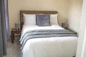 A neatly made bed with a white comforter and two pillows, flanked by two bedside tables with lamps, in a simple room with a plain wall and a curtain on one side.