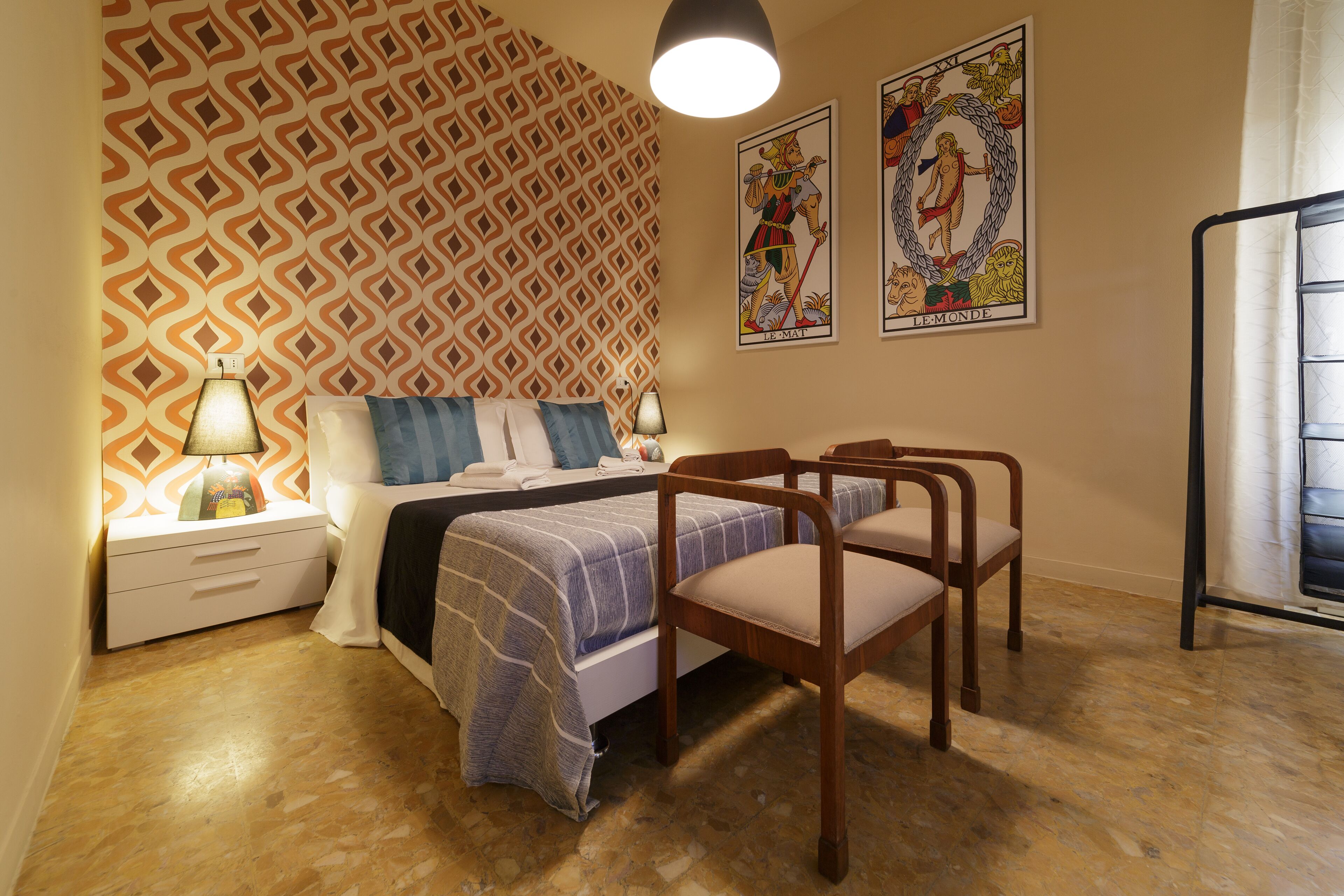 A bedroom with a patterned wallpaper, a bed with a blue and white bedspread, two wooden chairs, a nightstand with a lamp, and two posters on the wall.