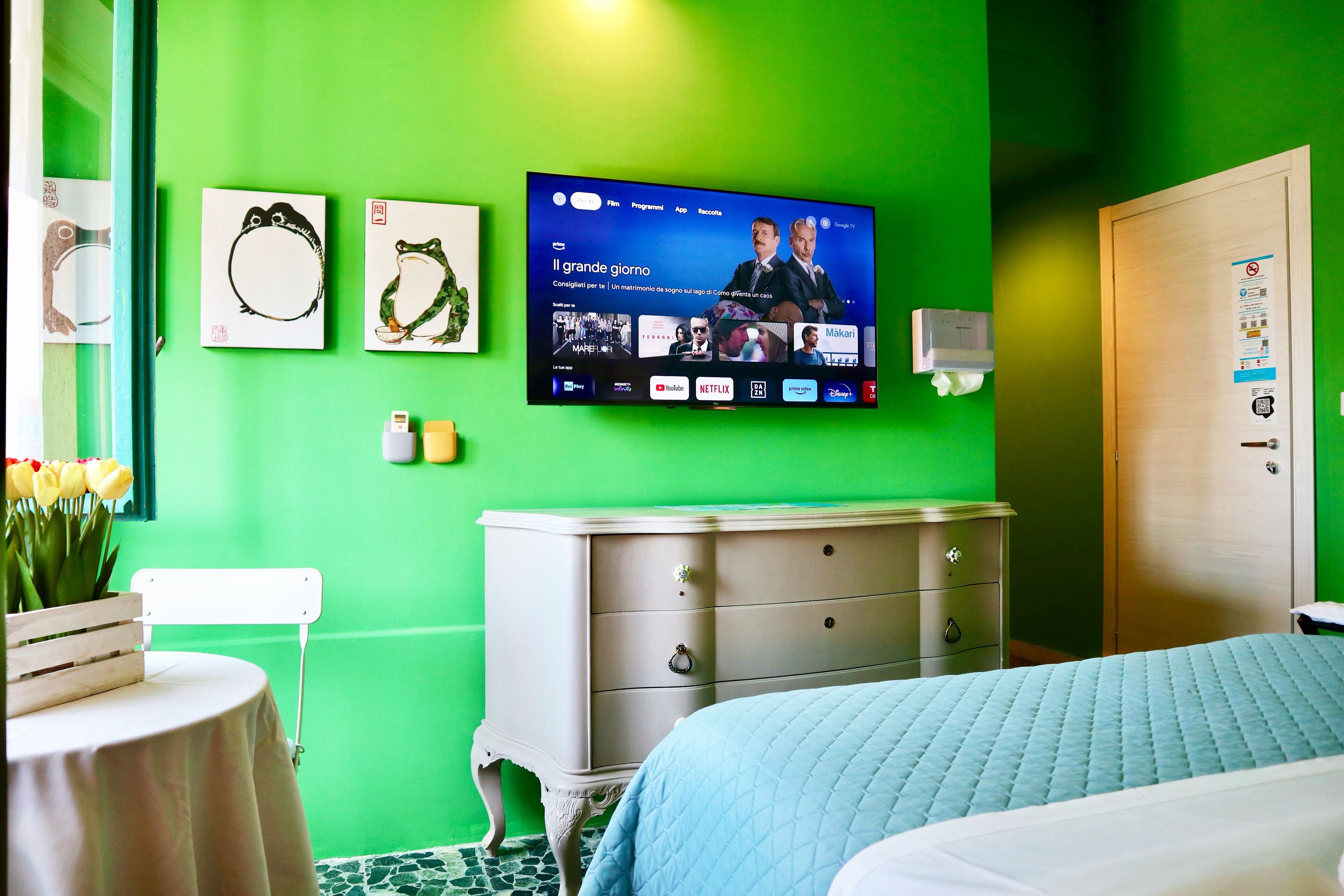 Quarto de hotel com parede verde, televisão de tela plana montada sobre um penteou branco, cama com colcha azul e uma pequena mesa com um vaso de flores.