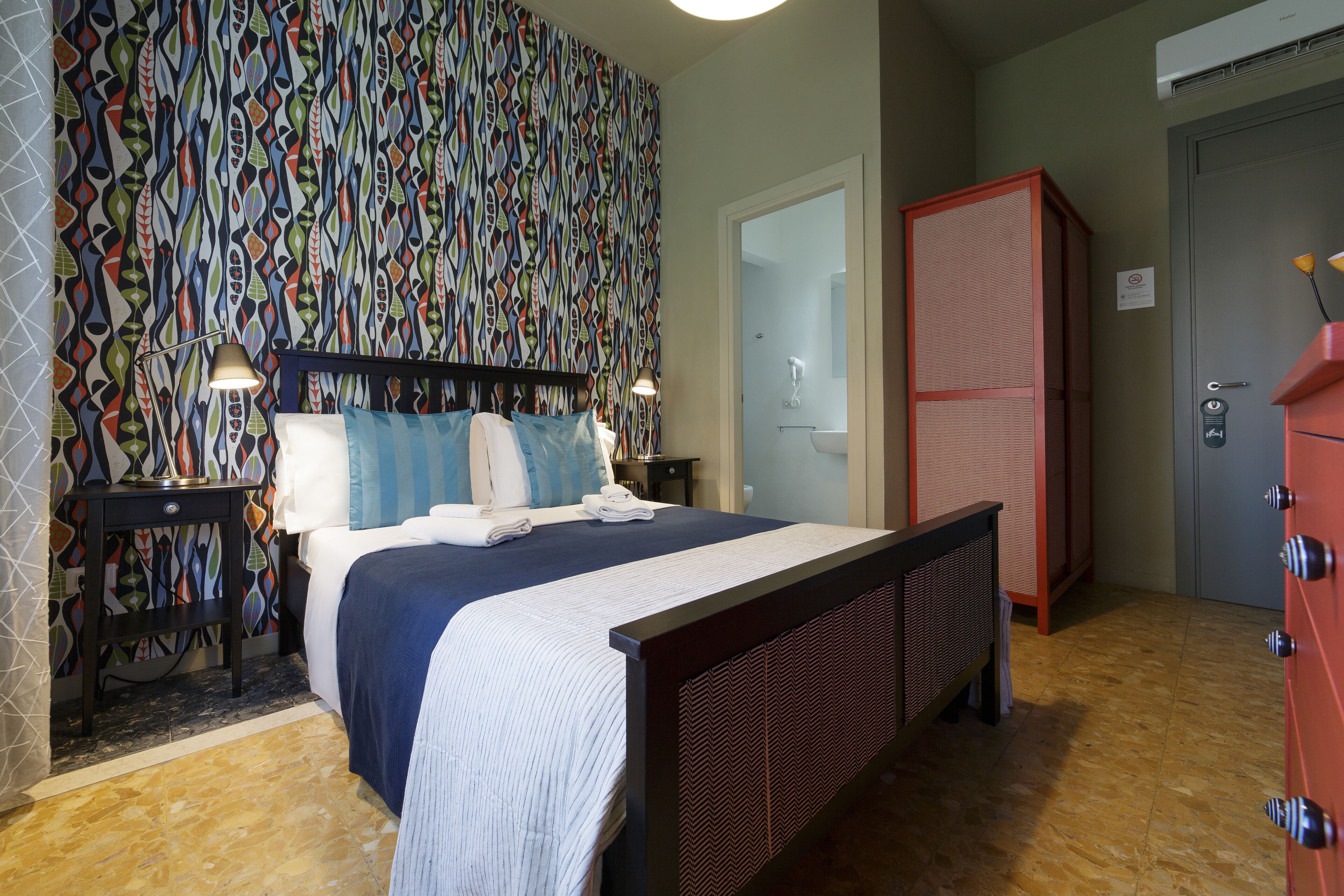 Une chambre à coucher avec un papier peint à motifs, un lit recouvert d’une couvre-lit bleue et blanche, deux lampes de chevet, une armoire et une porte sur laquelle est fixé un porte-serviettes.