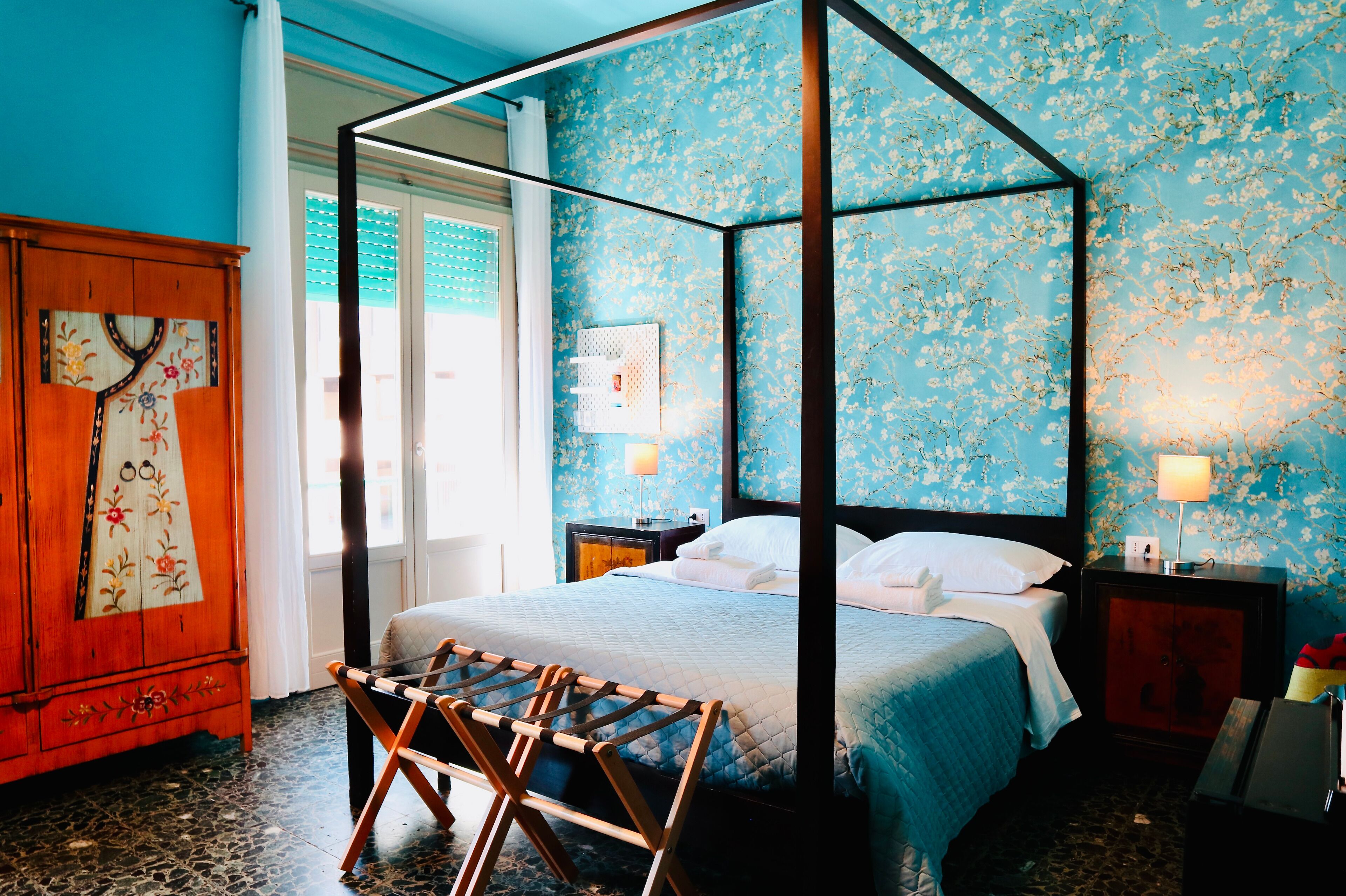 A four-poster bed with a blue floral wallpaper, a wooden cabinet with floral embroidery, and a small wooden stool.