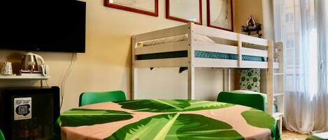 A room with a bunk bed, a table with a green and pink tablecloth, a television mounted on the wall, and a small refrigerator.