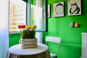 Una stanza con una parete verde, una finestra che si affaccia su edifici, un tavolo con una tovaglia bianca, una cassa di legno con fiori e opere d'arte appese al muro.