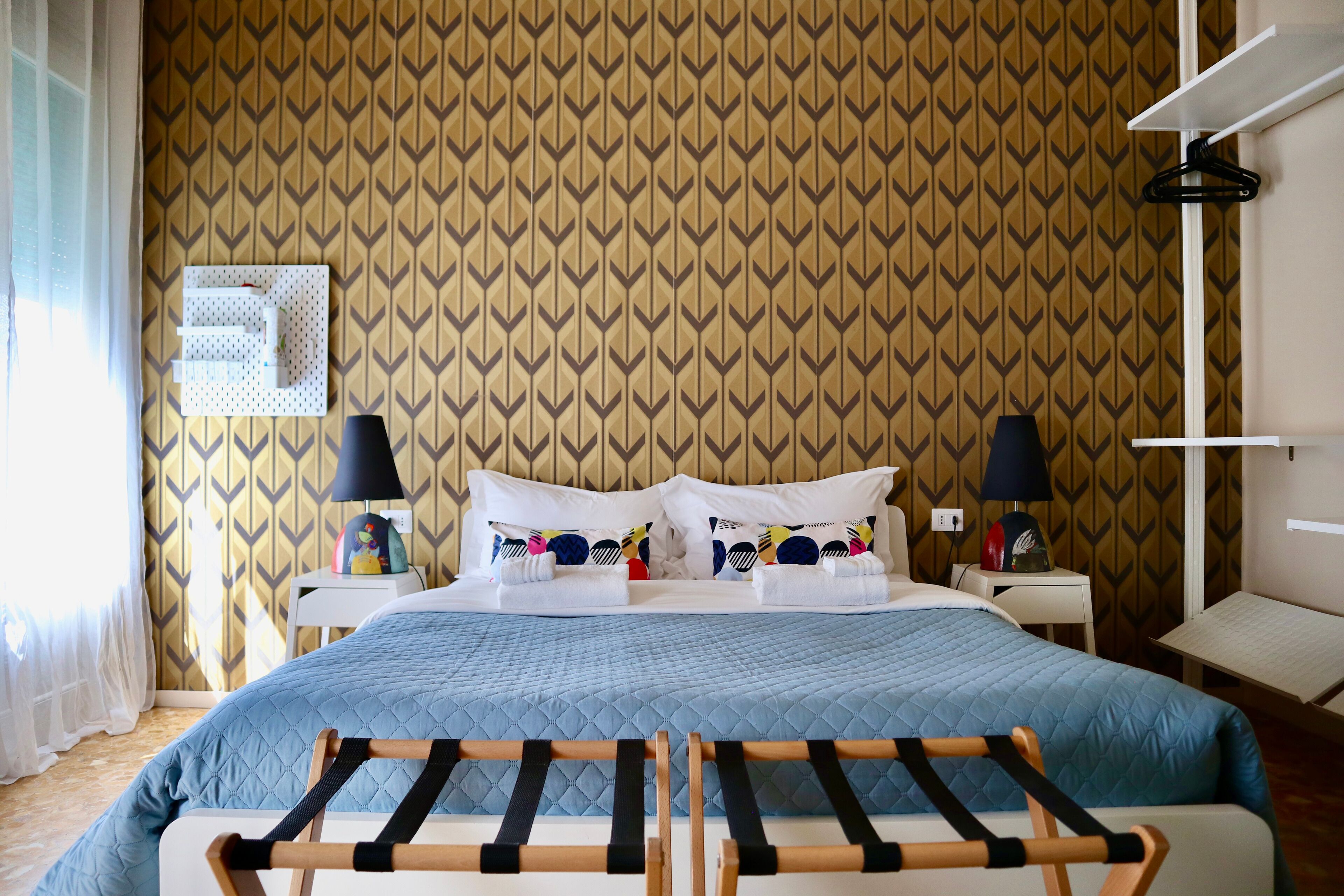 A hotel room with a patterned wallpaper, a bed with blue bedding, two bedside tables with lamps, and a wall-mounted shelf.