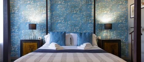 Un lit à baldaquin avec un papier peint à motifs floraux bleus, deux tables de chevet avec des lampes et une vue sur l’architecture de la pièce.