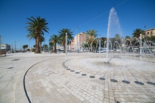 Una piazza pubblica con una fontana al centro, palme e edifici sullo sfondo.