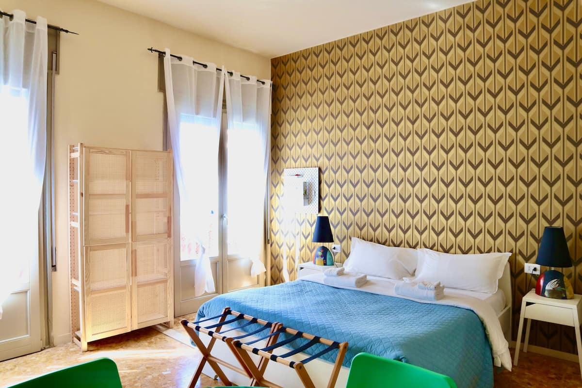 Una habitación de hotel con cama, mesita de noche, lámpara y ventana con cortinas.