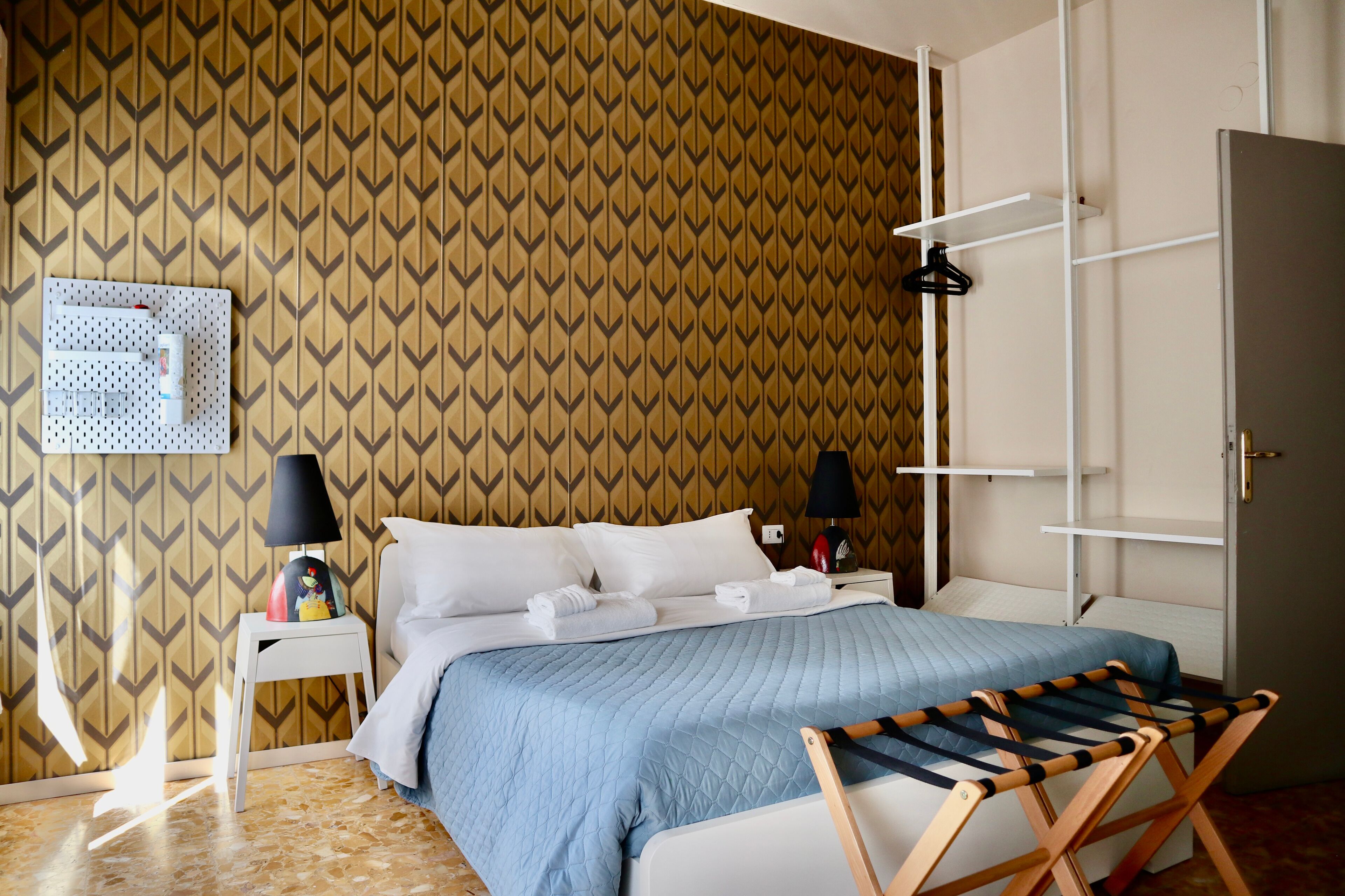 Une chambre d’hôtel avec un papier peint à motifs, un lit recouvert d’une courtepointe bleue, deux tables de chevet avec des lampes, un calendrier mural et une chaise en bois.