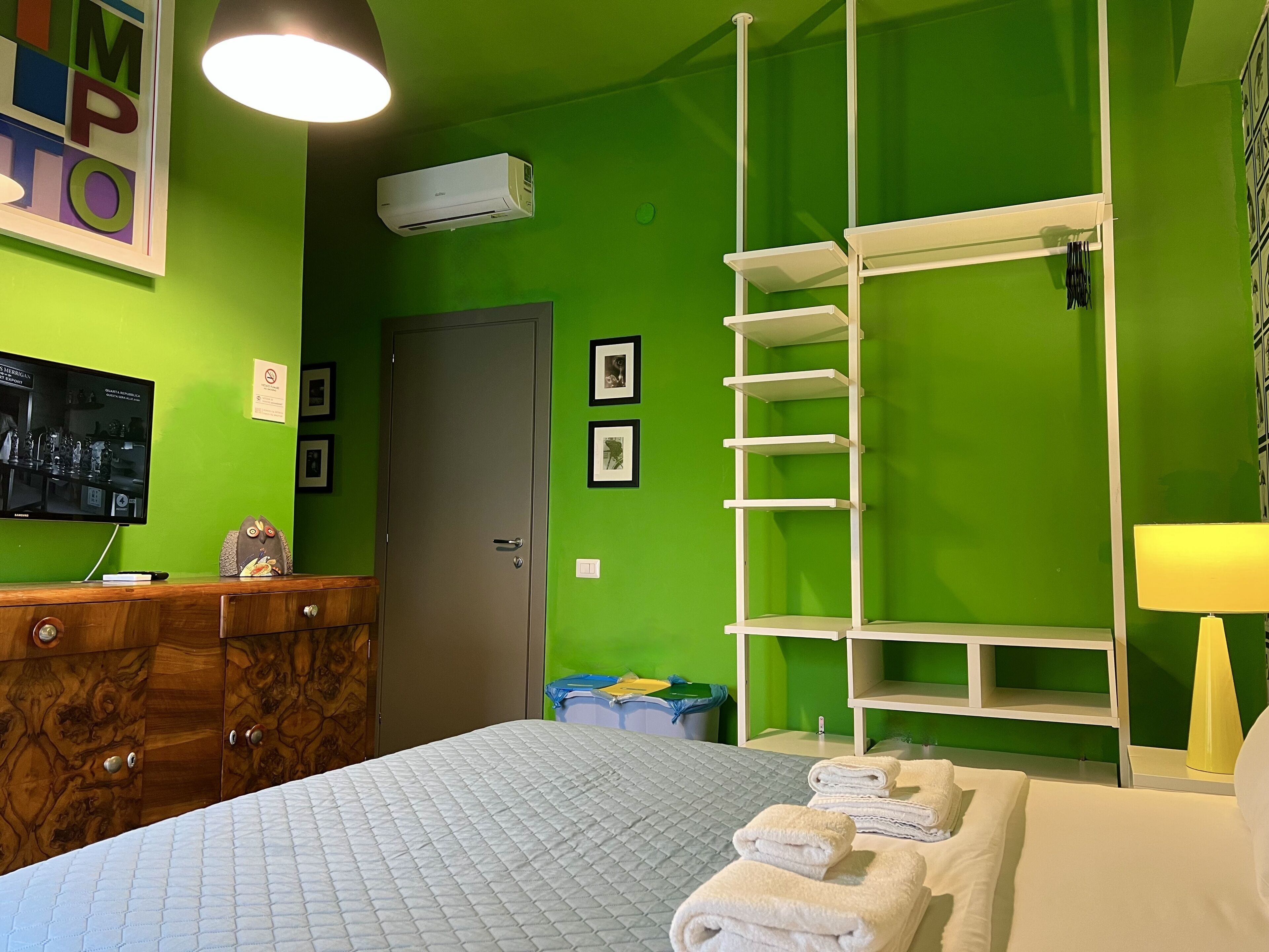 Een hotelkamer met een groene wand, een bed met een gewatteerde spreidek, een witte plankkast en een wandairconditioning.