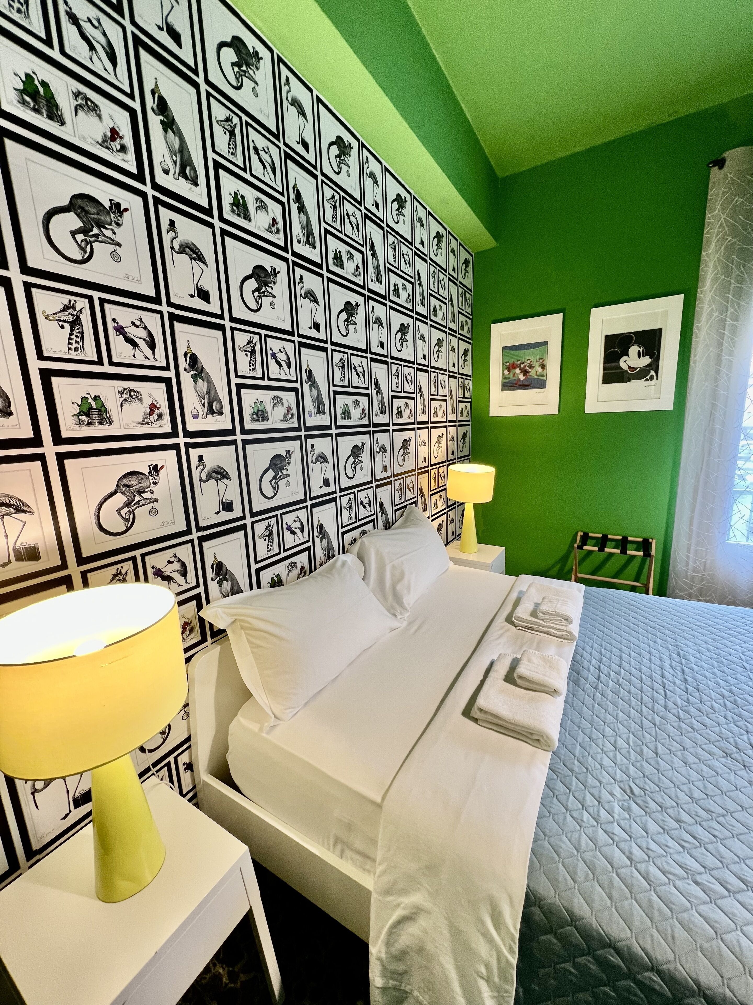 A hotel room with a green wall, a bed with white linens, a yellow lamp, and framed pictures on the wall.