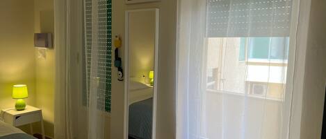 A hotel room with a bed, a bedside table with a lamp, a mirror, and a window with sheer curtains.