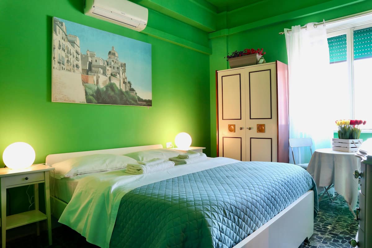 Una cama bien hecha con colcha verde, una lámpara de noche y un cuadro de un pueblo costero en la pared.