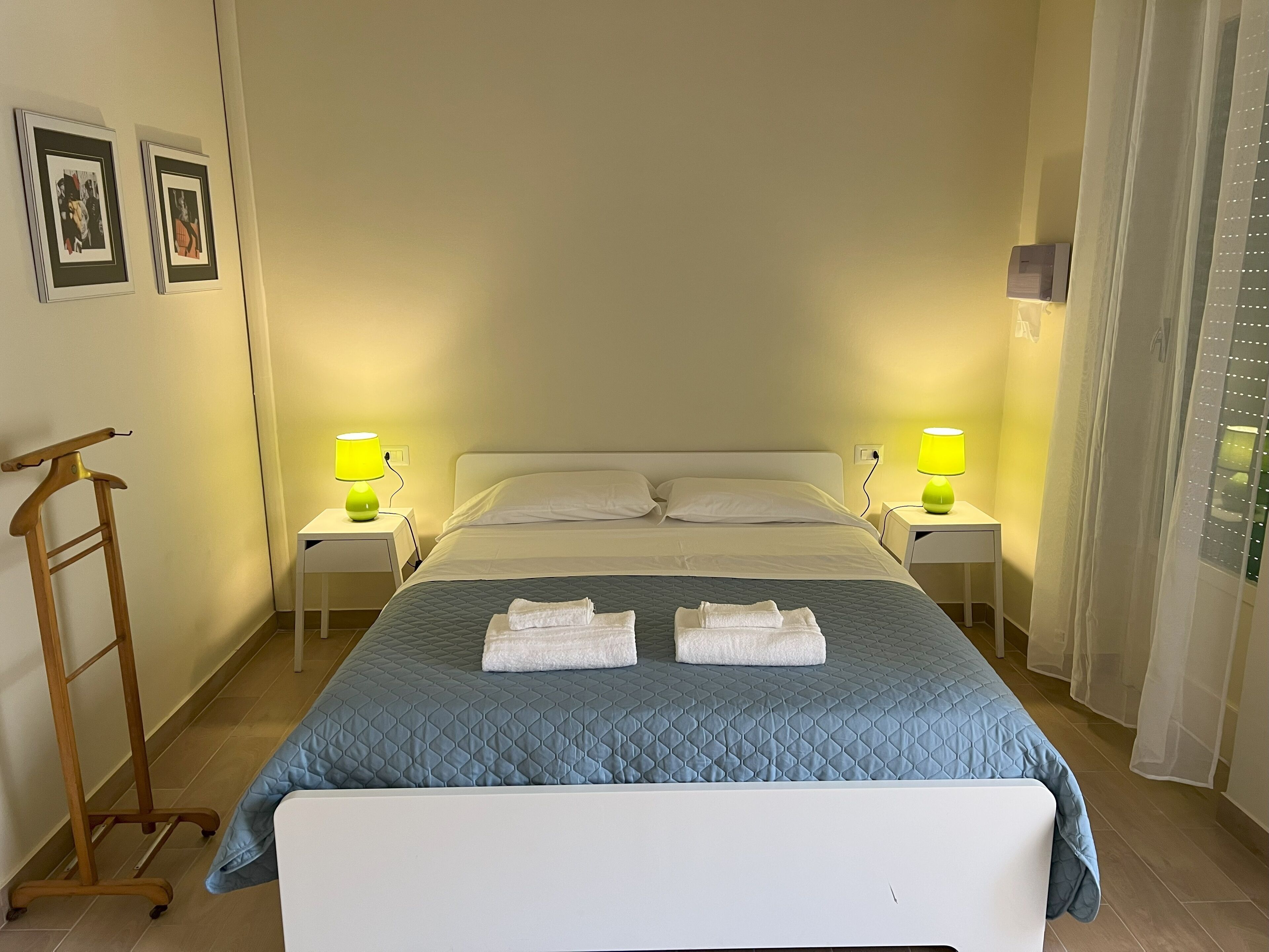A neatly made bed with a blue quilt, two white pillows, and two folded towels on the bed. There are two bedside tables with lamps, a wooden hanger on the floor, and framed pictures on the wall.
